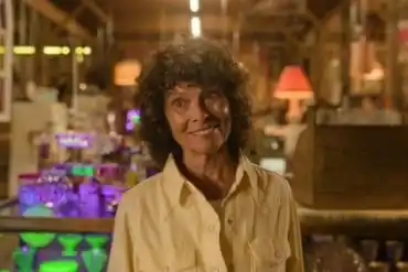 Elderly woman with curly hair smiling in a cozy, colorful store setting.