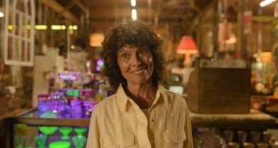Elderly woman with curly hair smiling in a cozy, colorful store setting.