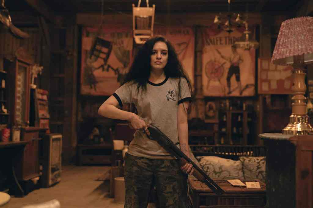Woman with long dark hair holding a shotgun in a dimly lit room.