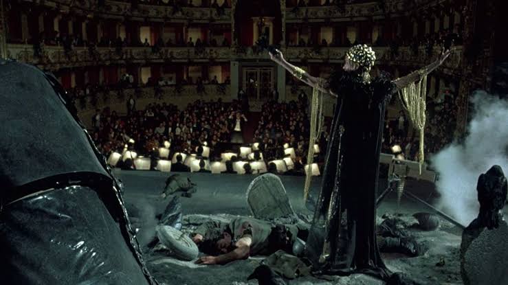 Dark theatrical scene with a figure in black and a skeleton in a grand theater setting.