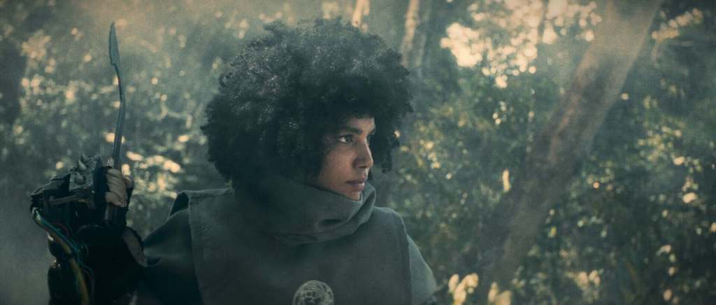 Young woman with curly hair in a forest, holding a bow and arrow, focused and alert.