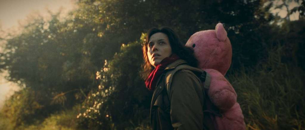 Woman with a pink teddy bear backpack in a forest at sunset.