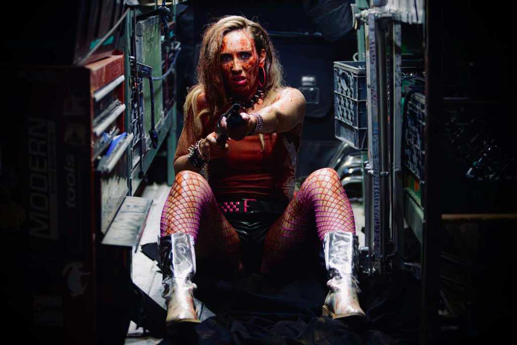 Woman with bloodied face and gun in a dark, cluttered room.