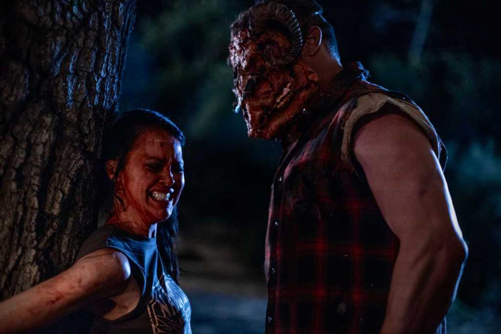 Scary creature with a mask confronts a woman in a dark outdoor setting.
