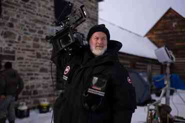 Cameraman in winter gear filming outdoors in snowy environment.